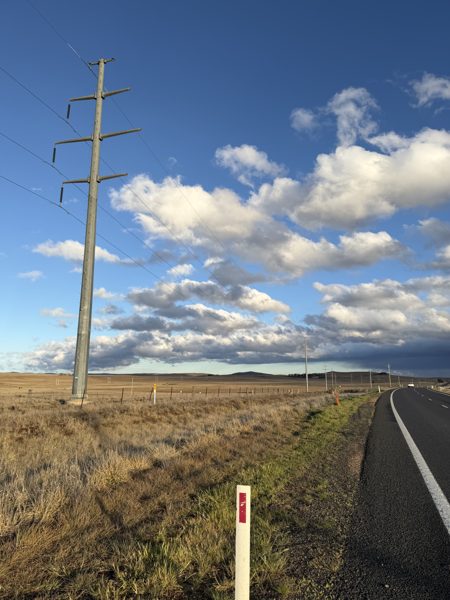 A galvanised steel 132 kilovolt pole beside the Monaro Highway under a sky of scattered cloud, with open country to the horizon.