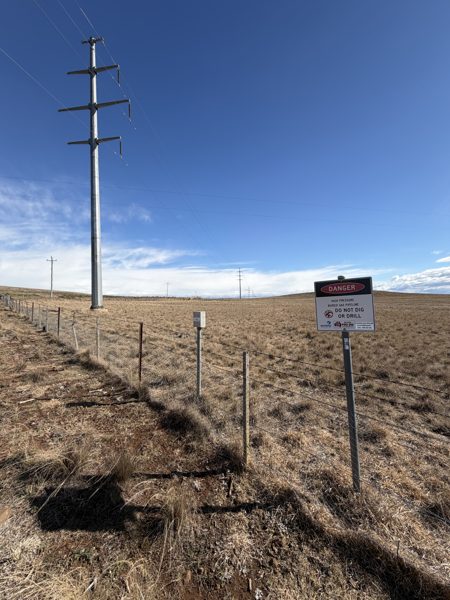 A tall galvanised steel 132 kilovolt pole with three cross-arms beside a farm fence, and a high pressure gas pipeline danger sign a few metres along the same fence.