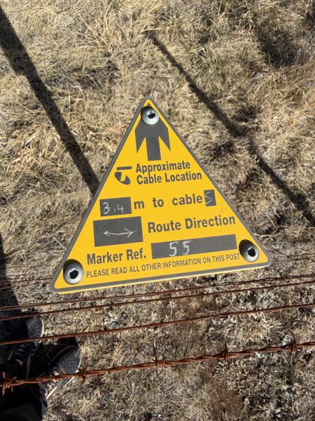 A yellow triangular Telstra cable marker on a fence post reading approximate cable location, 3.4 metres to cable, route direction along the fence, marker reference 55.