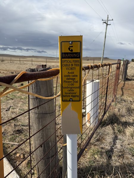 A yellow Telstra warning post fixed to a farm gate reading Warning, optical fibre cable in this vicinity, with open grazing land and a timber power pole behind.