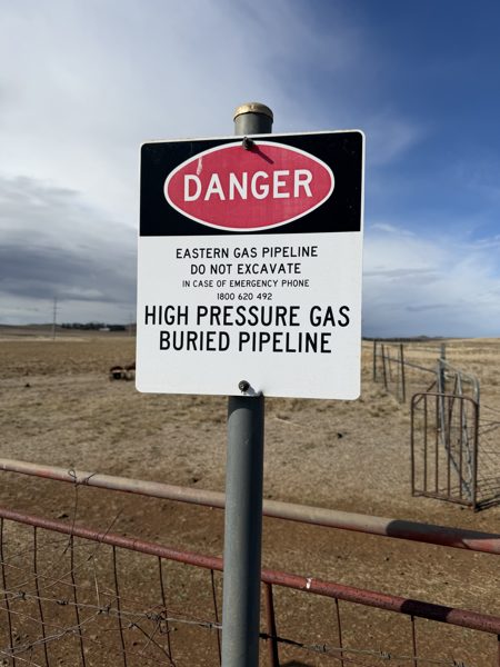 A white and red Danger sign on a steel post reading Eastern Gas Pipeline, do not excavate, high pressure gas buried pipeline, with open paddocks behind.