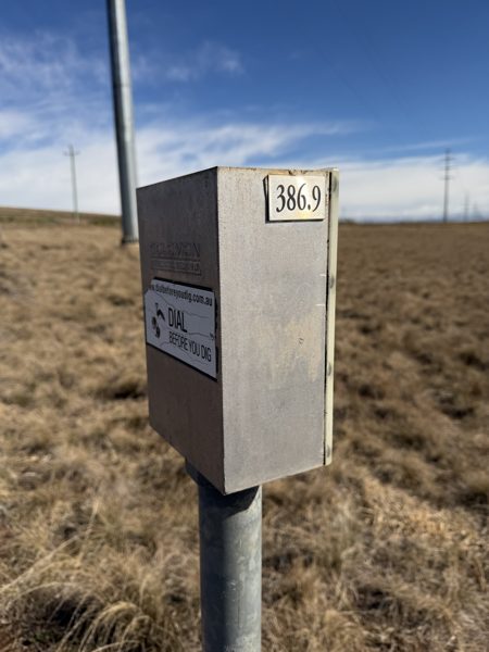 A galvanised marker post with a metal box reading 386.9 and a Dial Before You Dig sticker, with the 132 kilovolt pole line on the horizon.