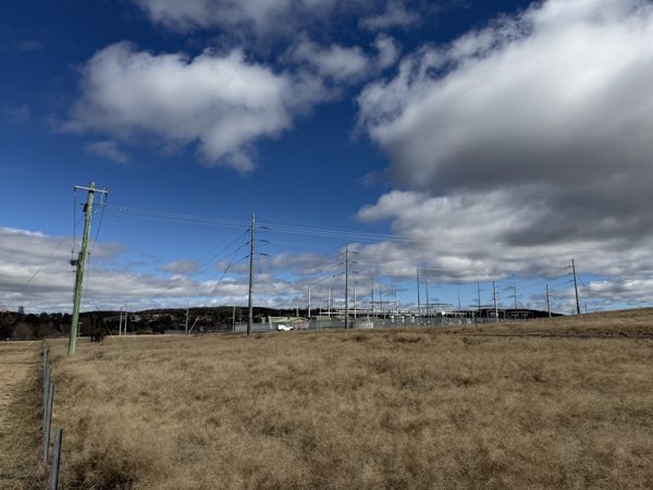 A fenced electricity substation with gantries, busbars and multiple incoming pole lines, seen across dry grassland on the edge of a town.